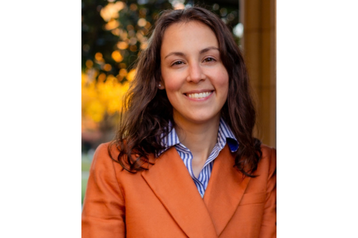 Professional headshot of a smiling woman with long, wavy dark brown hair. She wears a burnt-orange blazer over a blue-and-white striped shirt and stands outdoors in front of softly blurred autumn foliage in golden yellow tones.
