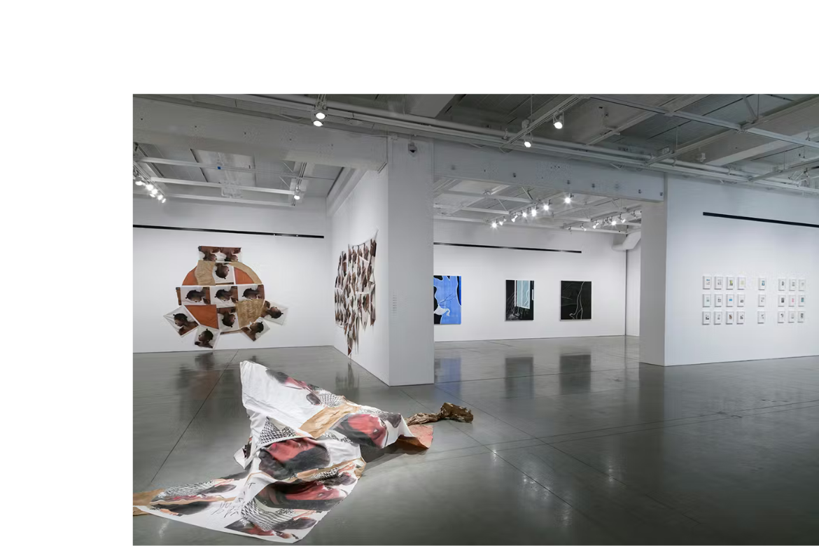 A spacious contemporary art gallery with polished concrete floors and white walls displays a mix of large-scale mixed-media installations, hanging artworks, and framed pieces under bright overhead lighting. In the foreground, a crumpled fabric-like artwork lies across the floor, while additional abstract and collage-style works are mounted on walls throughout the open exhibition space.